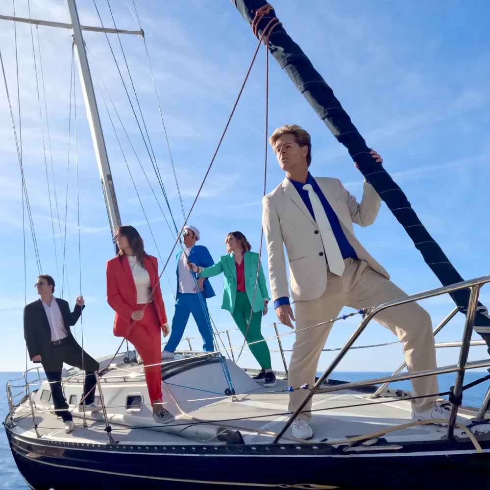 New Moon on Monday band members in bright suits standing on a sailboat deck under a blue sky